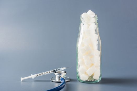 Glass bottle full of white sugar cube sweet food ingredient and doctor stethoscope isolated gray background, health high blood risk of diabetes and calorie intake concept and unhealthy drink