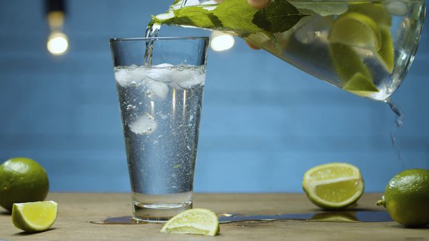 Close up pouring lemonade with lime and mint into a glass. Blurry lamps on blue background