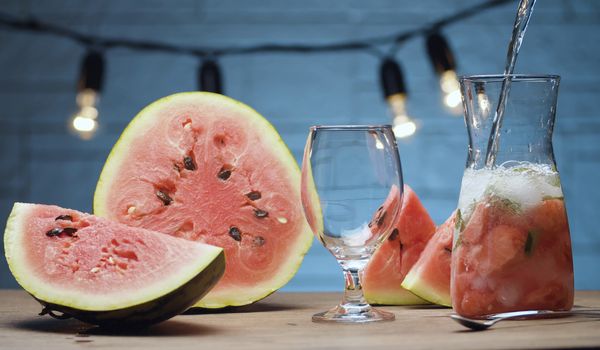 Close up preparing watermelon lemonade. Pouring soda water into a jug with pieces of watermelon, mint leaves and ice. Blurry lamps on blue background