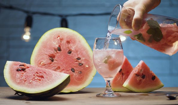 Close up male hand pouring lemonade into a glass from a jug with pieces of watermelon, mint leaves and ice. Blurry lamps on blue background