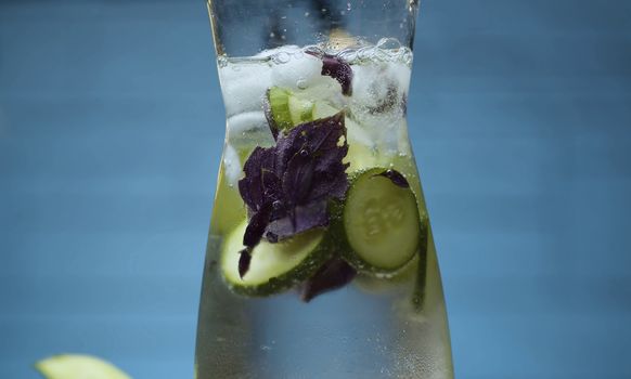 Close up vegetable fizzy drink. Sparkling water in a glass jug with sliced cucumber and purple basil leaves. Blurry bulbs on blue background