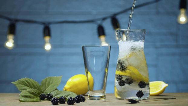 Close up cooking dewberry lemonade. Pouring soda water into a jug with dewberry, sliced lemon and ice. Blurry lamps on blue background