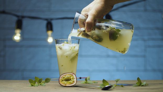 Close up male hand pouring drink with mint and passion fruit into a glass. Blurry lamps on blue background