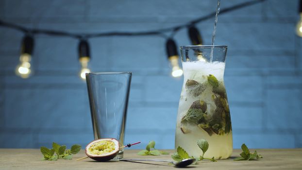 Close up pouring fizzy water in the jug with mint and passion fruit drink. Blurry lamps on blue background