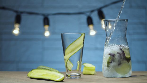 Close up cooking vegetable drink. Pouring fizzy water into a glass with sliced cucumber and purple basil leaves. Blurry bulbs on blue background