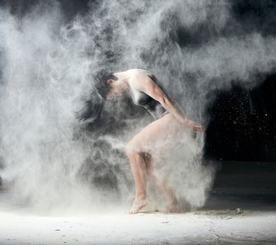 beautiful caucasian woman in a black bodysuit with a sports figure is dancing in a white cloud of flour on a black background