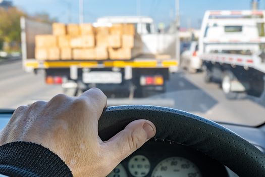 view of the driver hand on the steering wheel of a car that is in a traffic jam at a traffic light in front of a truck with a dangerous load of incorrectly fixed lumber, timber