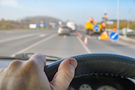 view of the driver hand on the steering wheel of a car against the background of road signs, construction equipment for road repairs, fences and passing cars in front