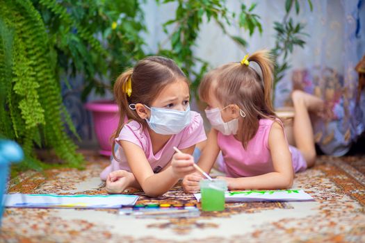 Two girls in protective medical masks paint with paints lying on the floor at home or in kindergarten. Children in self-isolation or quarantine