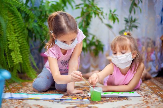 Two girls in protective medical masks paint with paints lying on the floor at home or in kindergarten. Children in self-isolation or quarantine