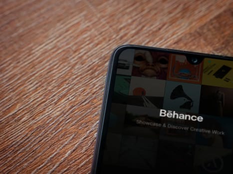 Lod, Israel - July 8, 2020: Adobe Behance app launch screen with logo on the display of a black mobile smartphone on wooden background. Top view flat lay with copy space. 