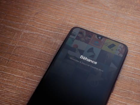 Lod, Israel - July 8, 2020: Adobe Behance app launch screen with logo on the display of a black mobile smartphone on wooden background. Top view flat lay with copy space. 