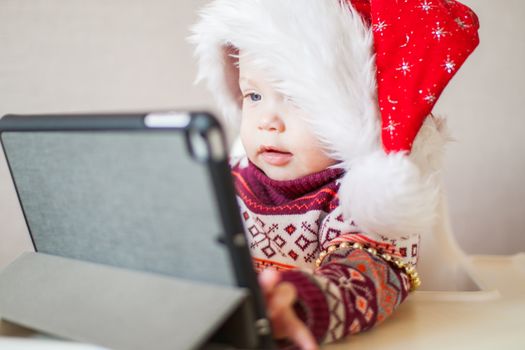 Little baby girl in Santa Claus hat looks at the tablet screen. Talk with family, Happy New Year and Merry Christmas.