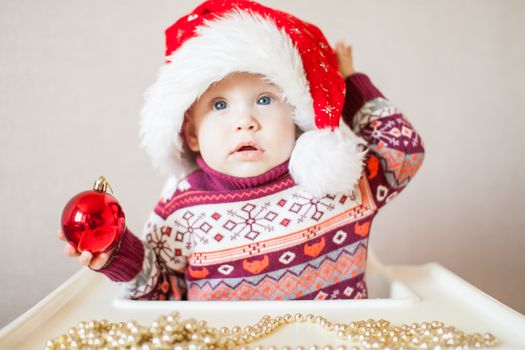 A little baby girl in a New Year's hat of Santa Claus examines and plays with New Year's decorations. Merry Christmas and Happy New Year greetings.