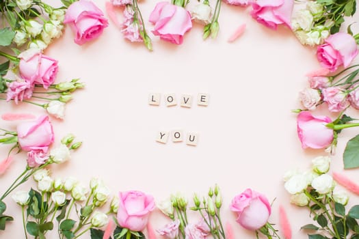 Inscription of wooden blocks love you. Frame of delicate white and pink roses and eustomas on a light pink background. Layout. Flat lay