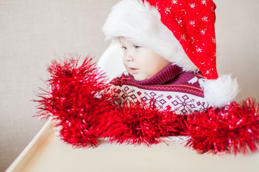 A little baby girl in a New Year's hat of Santa Claus examines and plays with New Year's decorations. Merry Christmas and Happy New Year greetings.