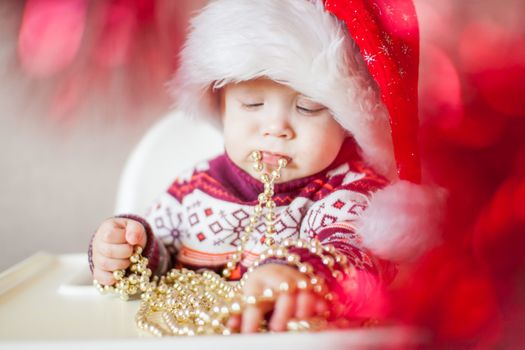 A little baby girl in a New Year's hat of Santa Claus examines and plays with New Year's decorations. Merry Christmas and Happy New Year greetings.