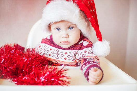 A little baby girl in a New Year's hat of Santa Claus examines and plays with New Year's decorations. Merry Christmas and Happy New Year greetings.