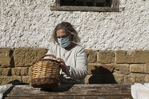 A senior adult woman, wearing a face mask, searches for something in a wicker basket before eating in front of the mountain hut of the fountain of Artica, in Luesia, Spain.