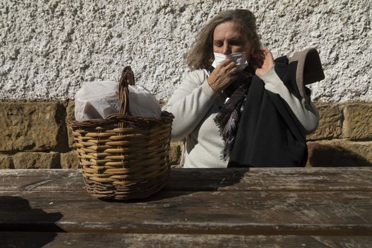 A senior woman, with short hair, sitting at the table in front of the mountain hut of the fountain of Artica, in Luesia, Spain, correctly adjusts her protective face mask.