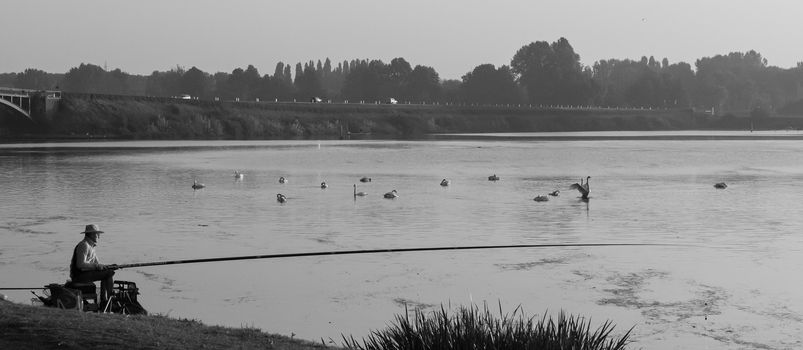 A fisherman with a long fishing rod sits on the river bank, black and white image