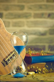A vintage hourglass and ukulele with an old book and brass pen on a wooden table and brick background in the morning. Closeup and copy space. The concept of memories or things in the past.