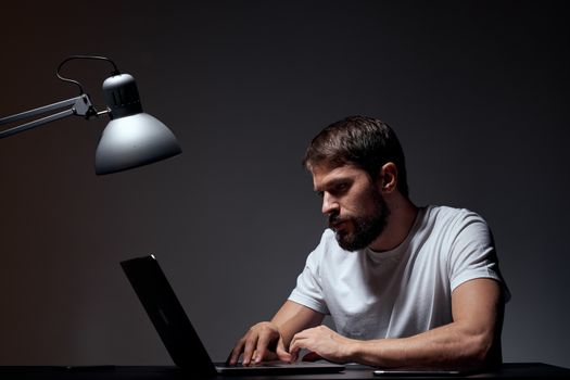 man with laptop sitting at table dark background workplace office emotions lamp gesturing with hands model cropped view. High quality photo