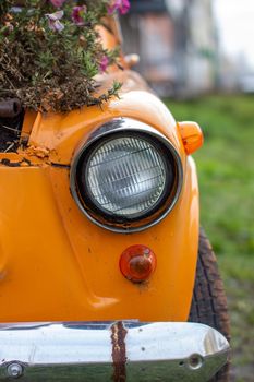 An old yellow car with round headlights and an open hood like a flower bed stands outside a flower shop in the city. Round headlight close-up