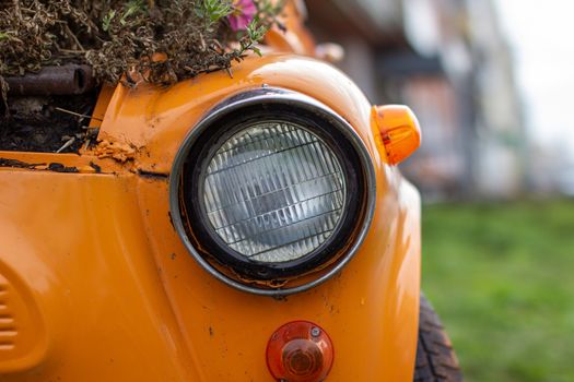 An old yellow car with round headlights and an open hood like a flower bed stands outside a flower shop in the city. Round headlight close-up