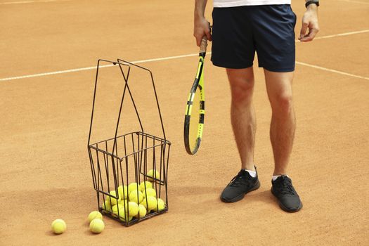 Man hold racket on clay court with basket of tennis balls