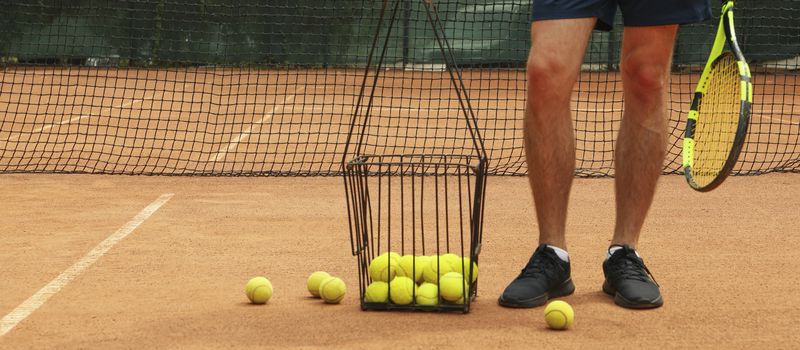 Man hold racket on clay court with basket of tennis balls