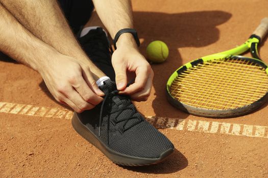 Man ties his shoelaces on clay court with racket and ball