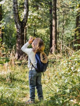 Curious boy is hiking in forest. Outdoor leisure activity for kids. Child looks through binoculars on tree foliage. Sunny day at autumn or summer day.