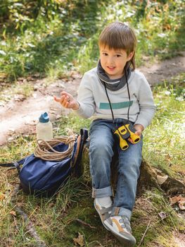 Little explorer on hike in forest. Boy with binoculars and compass sits on stump and reads map. Outdoor leisure activity for children. Summer journey for young tourist.
