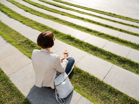 Woman with short haircut and eyeglasses sits in park with take away cup of coffee. Woman enjoys summer sun. Casual clothes, urban lifestyle of millennials.