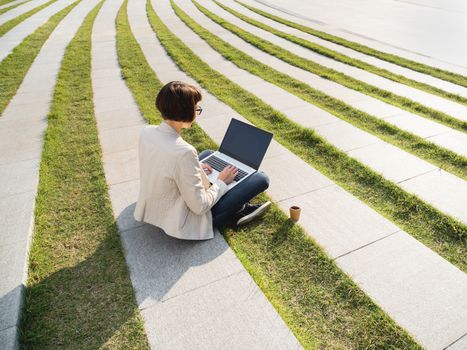 Freelance business woman sits in park with laptop and take away cardboard cup of coffee. Casual clothes, urban lifestyle of millennials. Working remotely.