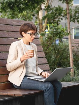 Freelance business woman sits in park with laptop and take away cardboard cup of coffee. Casual clothes, urban lifestyle of millennials. Working remotely.