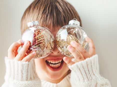 Kid with decorative balls for Christmas tree.Boy in cable-knit oversized sweater.Cozy outfit for snuggle weather.Transparent balls with red, golden spangles inside.Winter holiday spirit.New year.