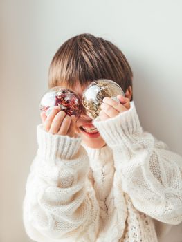 Kid with decorative balls for Christmas tree.Boy in cable-knit oversized sweater.Cozy outfit for snuggle weather.Transparent balls with red, golden spangles inside.Winter holiday spirit.New year.