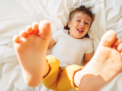 Little boy is lying upside down on the bed and laughing happily. Joyful toddler. Playful child shows his feet in the air. Sunny morning in cozy home.