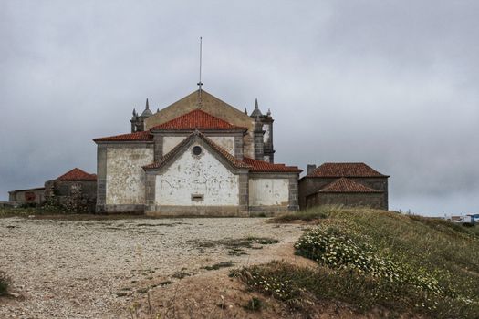 Sanctuary of Our Lady of Cape Espichel in Portugal under cloudy sky and surrounded by vegetation and wild colorful flowers