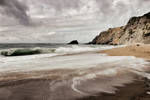 Brave ocean, rock formations and cloudy drama sky on the beach of Adraga in Sintra, Lisbon, Portugal