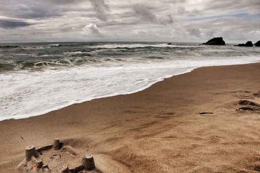 Brave ocean, rock formations and cloudy drama sky on the beach of Adraga in Sintra, Lisbon, Portugal. Sandcastle in the foreground.