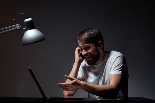 man with laptop sitting at table dark background workplace office emotions lamp gesturing with hands model cropped view. High quality photo