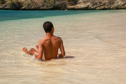 Rear view of a young man sitting on a sand beach. The blue sea and idyllic nature scene destination. Asian handsome teenager sitting and enjoying the view on the tropical beach.