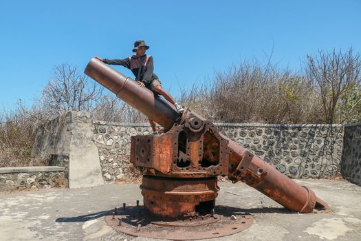 Asian teenager sits on a historic cannon. Military stone fortifications from World War II. A young guy sitting on an ancient cannon. Lombok, Indonesia.