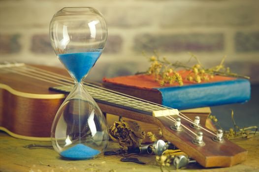 A vintage hourglass and ukulele with an old book and brass pen on a wooden table and brick background in the morning. Closeup and copy space. The concept of memories or things in the past.