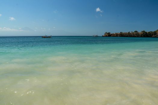 View of Rinjani volcano from Pink Beach Lombok. Bay with turquoise blue water and fishing boat anchored on calm sea level. Beach with its transparent water.