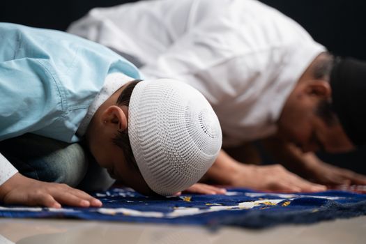 Muslim father and son praying or performing Salah while sitting on Prayer rug and touching head to mihrab or mosque printed on rug