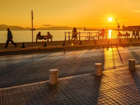 People along the promenade towards the city port at Nikis Avenue, during an orange sunset.